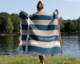 May include: A blue and tan striped throw blanket with fringed edges. The blanket has the text "THE WILSONS" and "PALM BEACH * EST. 2025" printed on it. The blanket is being held up by a person.