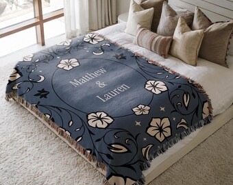 May include: A woven throw blanket in slate blue featuring a central oval frame with the text Matthew & Lauren. The design includes cream-colored floral motifs, dark swirling vines, and small star accents, finished with a fringed edge on all sides.