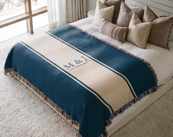 May include: A woven throw blanket featuring a deep blue color with a wide cream-colored central stripe. The text M & J is embroidered in the center of the stripe. The blanket has a fringed edge with multi-colored threads along the bottom border.