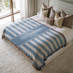 May include: A woven throw blanket featuring blue and cream stripes with a central diagonal band displaying the text The Sandersons est. 2026 in white script. The blanket has a fringed edge and is draped over a bed with several decorative pillows.