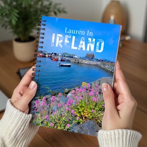 May include: A spiral-bound notebook with a vibrant cover featuring the text "Lauren In IRELAND" over a scenic coastal view. The image shows a harbor with boats, clear blue water, and pink flowers in the foreground. The notebook is held by a person.