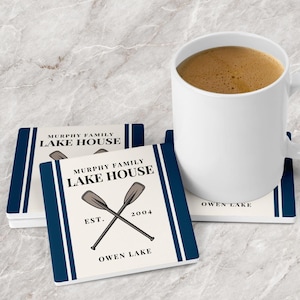 May include: Set of four white coasters with blue stripes and a navy blue border. The coasters feature a graphic of two crossed oars with the text "Murphy Family Lake House" and "Est. 2004" and "Owen Lake".
