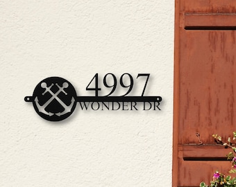 May include: A black metal house sign featuring a circular emblem with two crossed anchors on the left side. To the right, the numbers 4997 are displayed above the text WONDER DR, all integrated into a horizontal bar design with mounting holes.