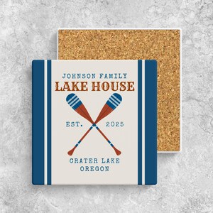 May include: A square coaster with a cork backing and a cream-colored top featuring two crossed oars in red and blue. The text reads JOHNSON FAMILY LAKE HOUSE, EST. 2025, and CRATER LAKE OREGON in blue and brown lettering with dark blue side borders.