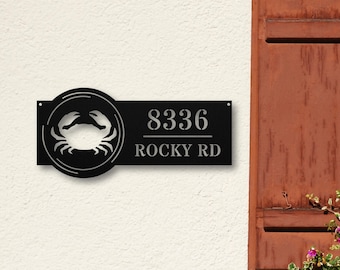 May include: A black metal address sign featuring a circular cutout of a crab on the left side. The right side displays the number 8336 above the text ROCKY RD in a clean, sans-serif font. The sign has small mounting holes on the far edges.