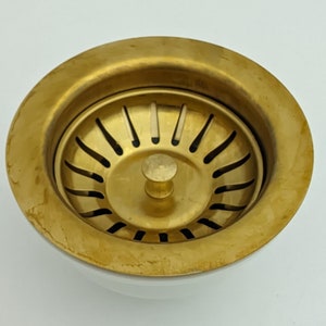 Könnte beinhalten: Ein goldfarbener Abflussstopfen aus Metall mit einem runden, flachen Boden und einem zentralen Abfluss mit einem kleinen Knopf. Der Abflussstopfen hat ein dekoratives, gemustertes Design.