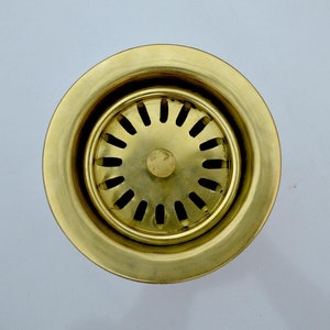 Puede incluir: Un colador de fregadero de metal de color dorado con un diseño circular y múltiples agujeros. El colador está diseñado para encajar sobre un desagüe de fregadero.