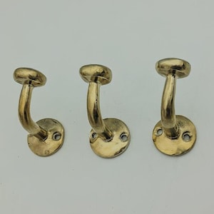 May include: Three gold-toned metal hooks with a curved design. The hooks are mounted on a white wall.