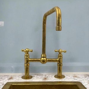 Pode incluir: Uma torneira de cozinha de latão com duas alças e um bico alto. A torneira está montada em uma bancada com um backsplash de azulejos brancos e marrons.