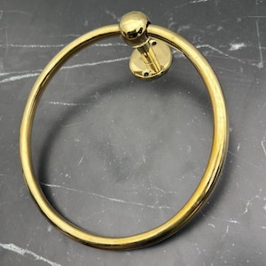 May include: A gold-toned metal towel ring with a round base and a smooth, circular design. The ring is attached to a wall.