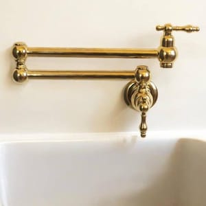 Pode incluir: Uma torneira dourada para encher panelas, montada na parede, com um design clássico. A torneira apresenta um braço de articulação dupla para estender o alcance, uma alça decorativa e um acabamento polido. Está instalada acima de uma pia branca.