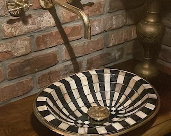 May include: A black and white patterned ceramic sink with a gold rim, mounted on a wooden countertop. The sink has a brass faucet with two handles. The countertop is in front of a brick wall with a gold mirror and a vase of dried flowers.