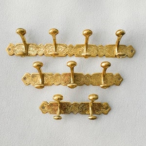 May include: A set of three gold-toned metal coat hooks with ornate designs. The hooks are attached to a decorative metal plate with a floral pattern.
