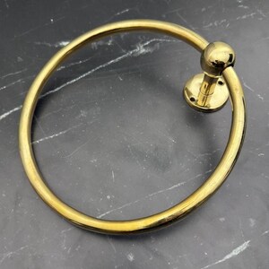 May include: A gold-toned metal towel ring with a round base and a smooth, circular ring. The ring is designed to hold towels or other items.