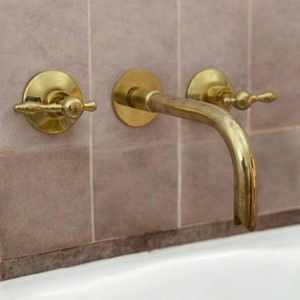 K&ouml;nnte beinhalten: Vergoldeter Badezimmerhahn mit zwei runden Griffen und einem gebogenen Auslauf, montiert an einer gekachelten Wand &uuml;ber einer wei&szlig;en Badewanne.