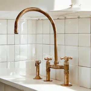 Può includere: Un rubinetto da cucina in ottone con un becco ad arco alto e due maniglie. Il rubinetto è montato su una parete piastrellata di bianco.