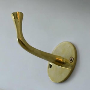 May include: A gold-toned metal hook with a curved design and a round base. The hook is attached to a wall with a screw.
