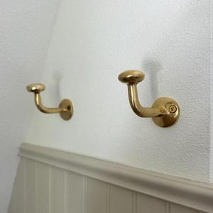 May include: Two brass-colored wall hooks with a curved design and rounded tops are mounted on a white wall. The hooks are attached with screws and are suitable for hanging items. The hooks are a decorative and functional addition to any space.