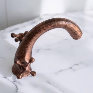Könnte beinhalten: Ein kupferfarbener Wasserhahn mit gebogenem Auslauf und kreuzförmigen Griffen. Der Wasserhahn weist auf dem Auslauf und der Basis aufwendige geätzte Muster auf, die einen dekorativen Akzent setzen. Geeignet für Badezimmer oder Küche.