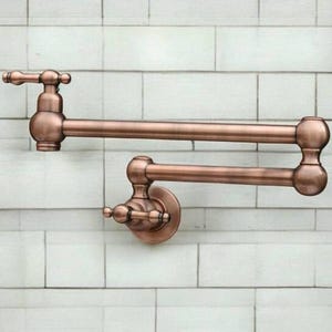 May include: A copper pot filler with a swivel spout and two arms. The pot filler is mounted on a white tiled wall.