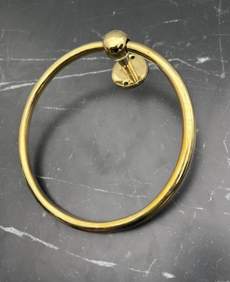 May include: A gold-toned metal towel ring with a round base and a smooth, circular design. The ring is attached to a wall.