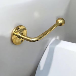 Puede incluir: Un porta rollo de papel higi&eacute;nico de metal dorado montado en una pared blanca. El soporte tiene una base redonda y un brazo curvo con un extremo redondo.