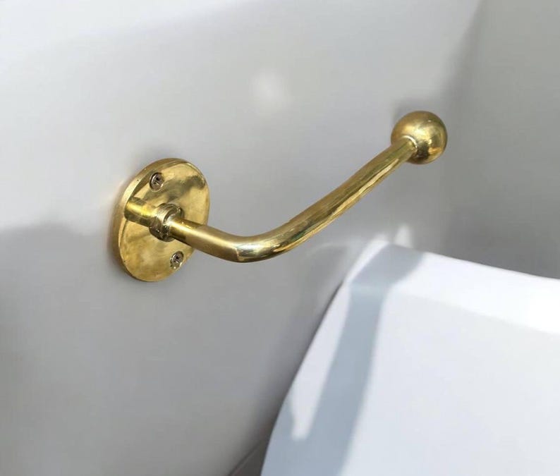 Puede incluir: Un porta rollo de papel higi&eacute;nico de metal dorado montado en una pared blanca. El soporte tiene una base redonda y un brazo curvo con un extremo redondo.