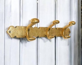 May include: A gold-colored metal coat rack with three curved hooks. The rack has a textured, dotted pattern and is mounted on a white surface. The hooks are evenly spaced and have a rounded design.
