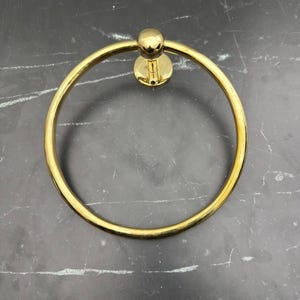 May include: A gold-toned metal towel ring with a round base. The ring is smooth and shiny.