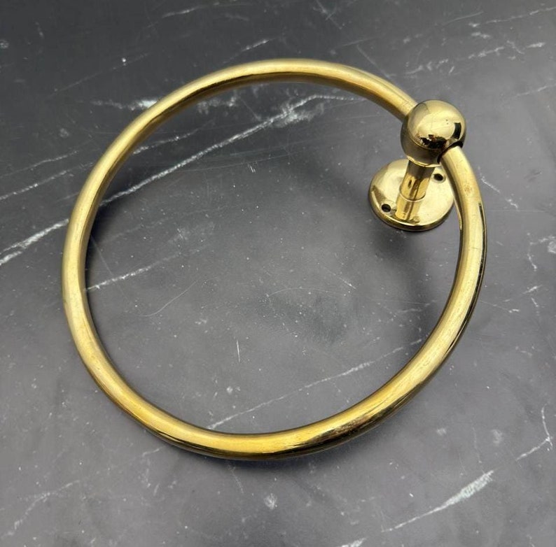 May include: A gold-toned metal towel ring with a round base and a smooth, circular ring. The ring is designed to hold towels or other items.