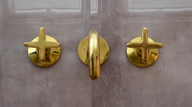 K&ouml;nnte beinhalten: Drei goldfarbene Badezimmerarmaturen mit kreuzf&ouml;rmigen Griffen. Die Armatur in der Mitte hat einen einzigen Griff, w&auml;hrend die beiden an den Seiten separate Griffe haben.