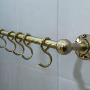 Peut inclure: Un porte-ustensiles de cuisine en métal doré avec cinq crochets et un bouton rond. Le porte-ustensiles est fixé à un mur en carrelage blanc.