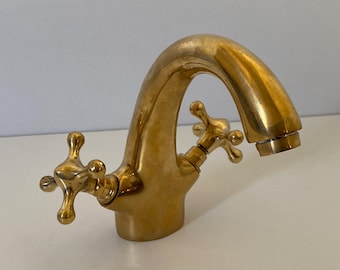May include: A gold-toned faucet with two cross-shaped handles. The faucet has a curved spout and a polished finish.