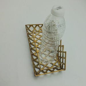 May include: A gold metal tray with a geometric design holds a clear plastic water bottle.