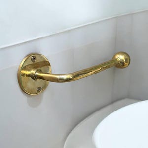 Puede incluir: Un porta rollo de papel higi&eacute;nico de metal dorado con una base redonda y un brazo curvo. El soporte est&aacute; montado en una pared blanca.