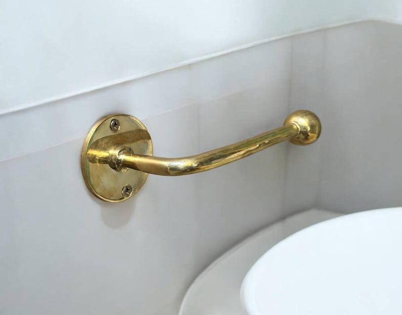 Puede incluir: Un porta rollo de papel higi&eacute;nico de metal dorado con una base redonda y un brazo curvo. El soporte est&aacute; montado en una pared blanca.