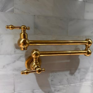 Peut inclure: Un robinet de remplissage de pot mural doré. Le robinet a un bras à double articulation et deux poignées avec des détails décoratifs. Il est installé sur un mur de carreaux de marbre blanc et gris.