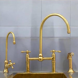 Könnte beinhalten: Ein goldfarbenes Küchenarmaturen-Set mit hohem Auslauf, Seitensprüher und separater Wasserfilterarmatur. Die Armaturen sind vor einer hellblauen gefliesten Rückwand angebracht. Der Wasserhahn hat zwei Griffe und ein klassisches Design.
