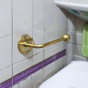 Puede incluir: Un porta rollo de papel higi&eacute;nico dorado montado en una pared de azulejos blancos con lechada morada. El soporte tiene una base redonda y un brazo curvo.