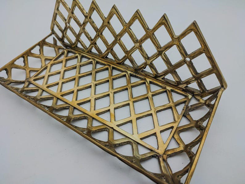 May include: A gold metal decorative stand with a diamond-shaped grid pattern. The stand is designed to hold items like books or magazines.