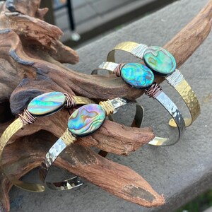 May include: Three gold and silver bracelets with abalone shell accents. The bracelets are stacked on a piece of driftwood.