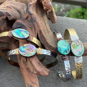 May include: Three bracelets with abalone shell accents. The bracelets are made of hammered metal with copper wire accents. The abalone shells are iridescent with shades of green, blue, and purple.
