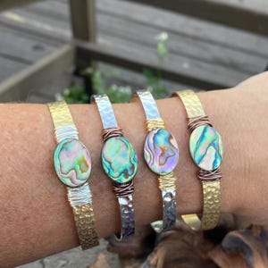 May include: Four hammered metal bracelets with abalone shell accents. The bracelets are in gold, silver, and copper tones.