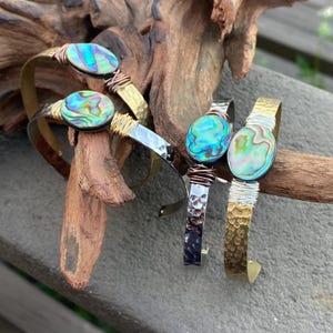 May include: Three bracelets with hammered metal bands and abalone shell accents. The bracelets are in gold, silver, and copper tones.
