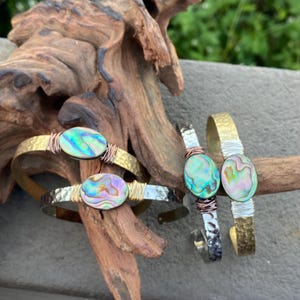 May include: Three bracelets with abalone shell accents. The bracelets are made of hammered metal with copper wire accents. The abalone shells are iridescent with shades of green, blue, and purple.