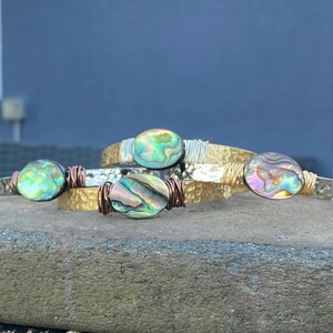 May include: Three gold-toned bracelets with abalone shell accents. The bracelets are stacked on top of each other and have a hammered texture.