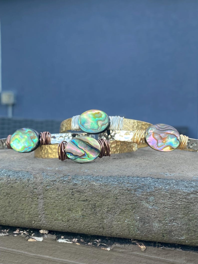 May include: Three gold-toned bracelets with abalone shell accents. The bracelets are stacked on top of each other and have a hammered texture.