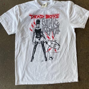 以下が含まれることがあります： 白いTシャツ。赤い文字で「DEAD BOYS」と書かれ、その下にマスクをした人物の白黒グラフィックと「SONIC REDUCER」の文字。グラフィックには稲妻とバナーが含まれています。