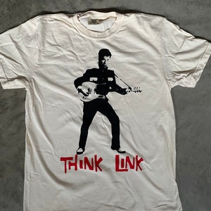 Puede incluir: Camiseta blanca con una silueta negra de una persona tocando una guitarra. El texto "THINK LINK" está impreso en rojo debajo de la imagen.