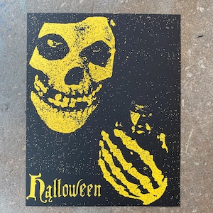 May include: Black and yellow Halloween-themed print featuring a skull and skeletal hand. The word "Halloween" is printed in a gothic font at the bottom. The skull and hand are yellow, and the background is black.